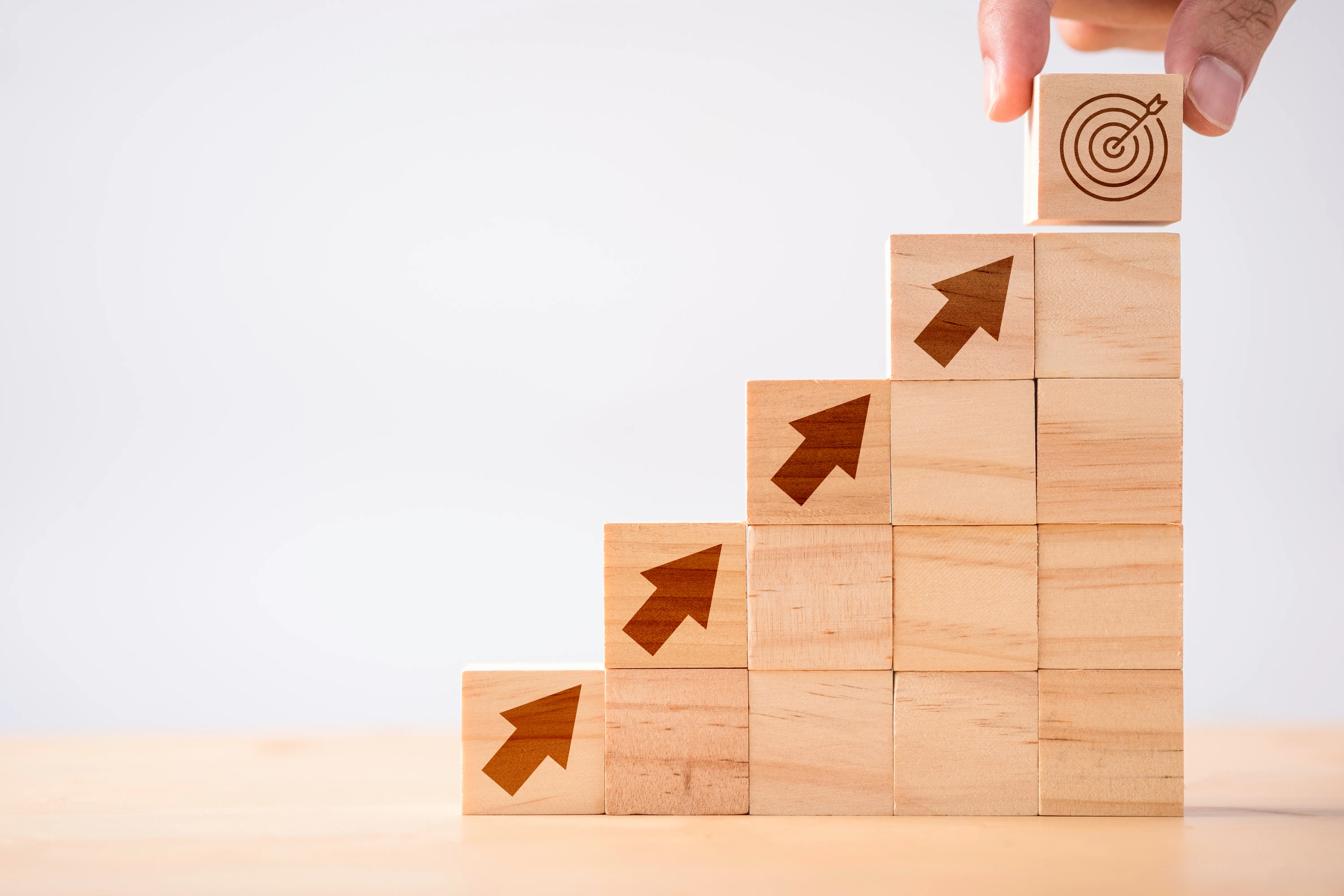 A hand placing a block marked with a target at the top of a rising staircase of wooden blocks